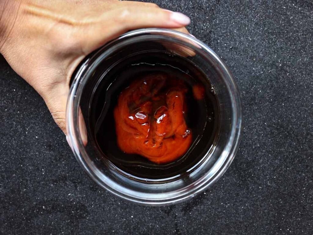 A hand holds a clear glass against a dark surface. Inside the glass is a dark liquid with a swirl of reddish sauce on top.